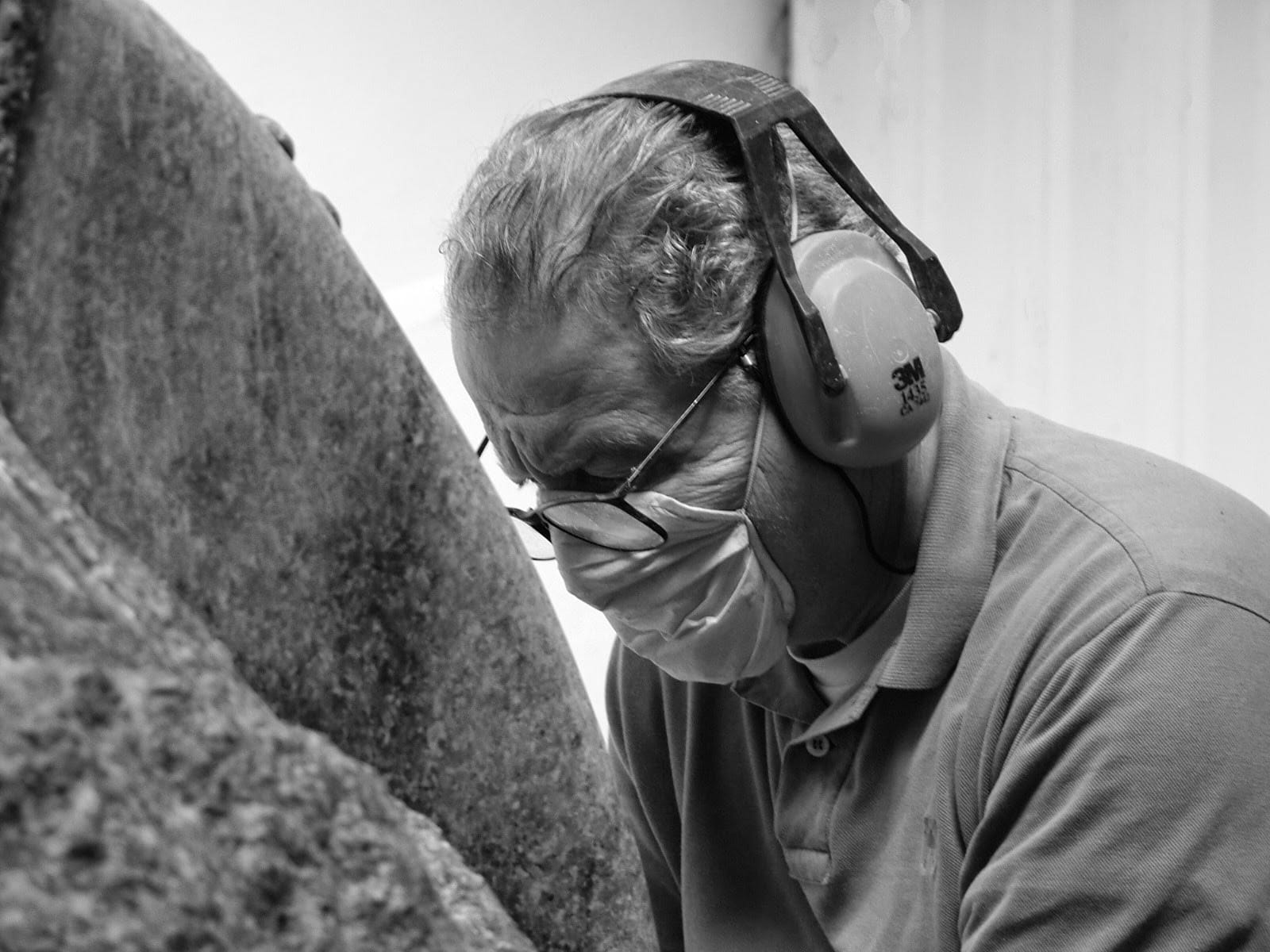 Imagem em preto e branco mostra o artista Paulo Aguinsky durante o processo de execução de sua arte. Ele tem os cabelos brancos, usa máscara cirúrgica, óculos de grau e fones profissionais de isolamento acústico.